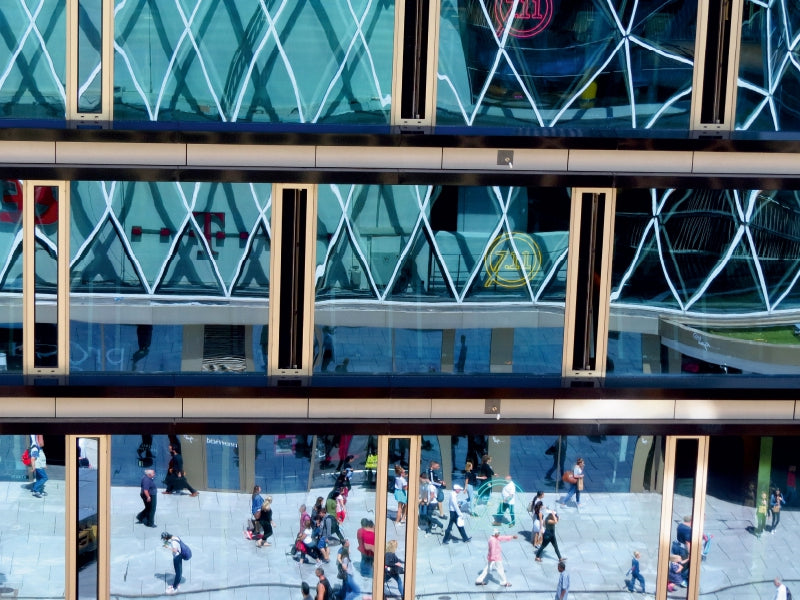 Moderne Glasfassade eines Gebäudes mit Spiegelungen von Fußgängern auf dem Gehweg.