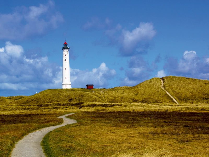 Ein hoher weißer Leuchtturm mit roter Spitze steht auf einer grasbewachsenen Düne unter blauem Himmel mit Wolken.