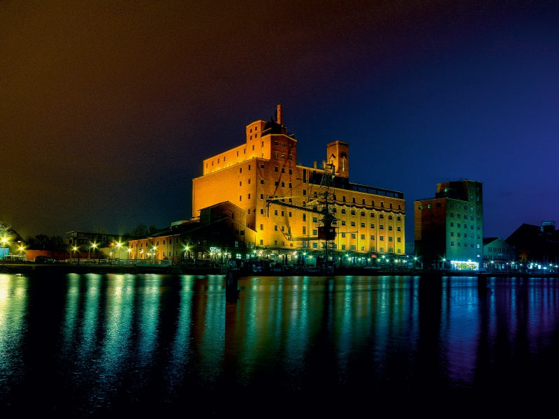 Beleuchtete Backsteingebäude bei Nacht spiegeln sich im dunklen Wasser.
