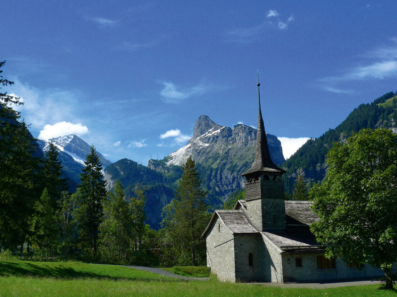 Eine Steinkirche mit spitzem Turm steht auf einer grünen Wiese vor hohen Alpenbergen und blauem Himmel.