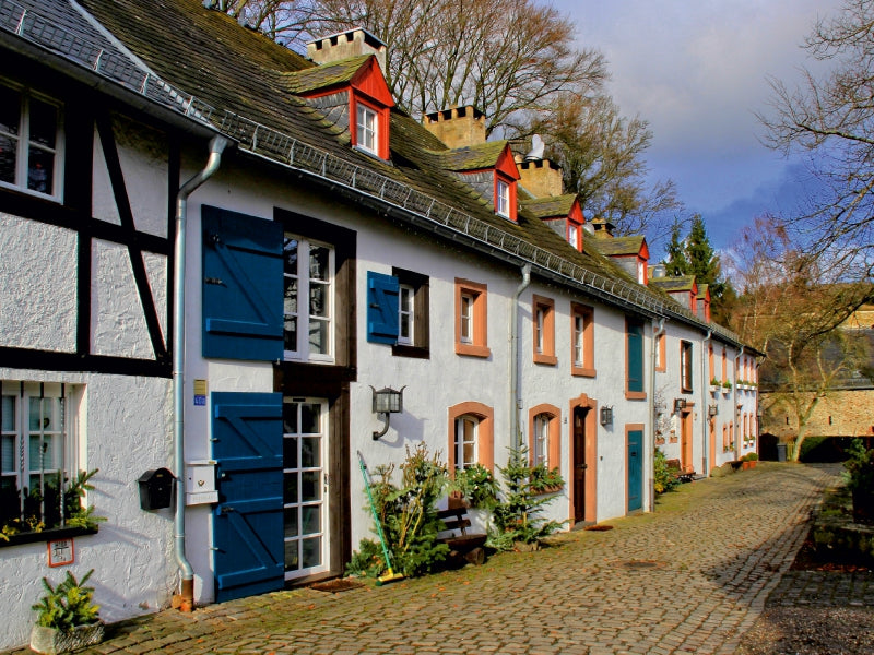 Eine Kopfsteinpflasterstraße führt an einer Reihe historischer Fachwerkhaeuser mit blauen Fensterlaeden vorbei.