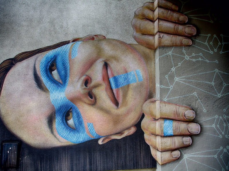 Wandmalerei eines lächelnden Gesichts mit blauer Bemalung und Händen, die um die Ecke schauen.