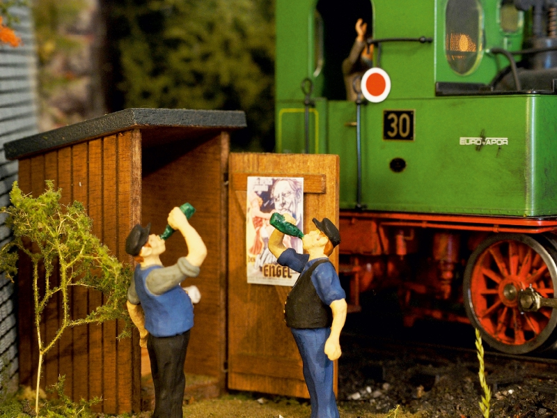 Modellfiguren trinken Bier vor einem Schuppen neben einer grünen Lokomotive.