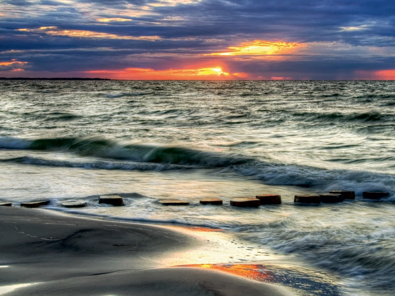 Sonnenuntergang am Meer mit sanften Wellen am Sandstrand und Holzbuhnen im Wasser.