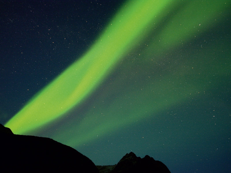 Grünes Nordlicht leuchtet über dunklen Bergen und einem sternenklaren Nachthimmel.