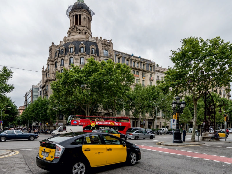 Ein belebter Straßenkreuzung in Barcelona mit Autos, einem roten Doppeldeckerbus und einem historischen Gebäude im Hintergrund.