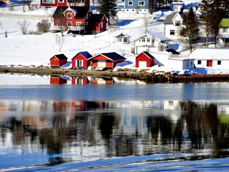 Schnee bedeckt eine malerische Küstenstadt mit roten und weißen Häusern, die sich im blauen Wasser spiegeln.
