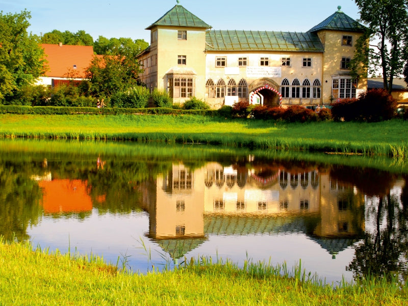 Ein historisches Schloss mit Türmen spiegelt sich im ruhigen Wasser eines Teiches, umgeben von grünem Gras und Bäumen.