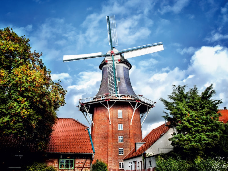 Eine große rote Backsteinmühle mit weißen Flügeln steht zwischen Bäumen unter einem blau-weißen Himmel.