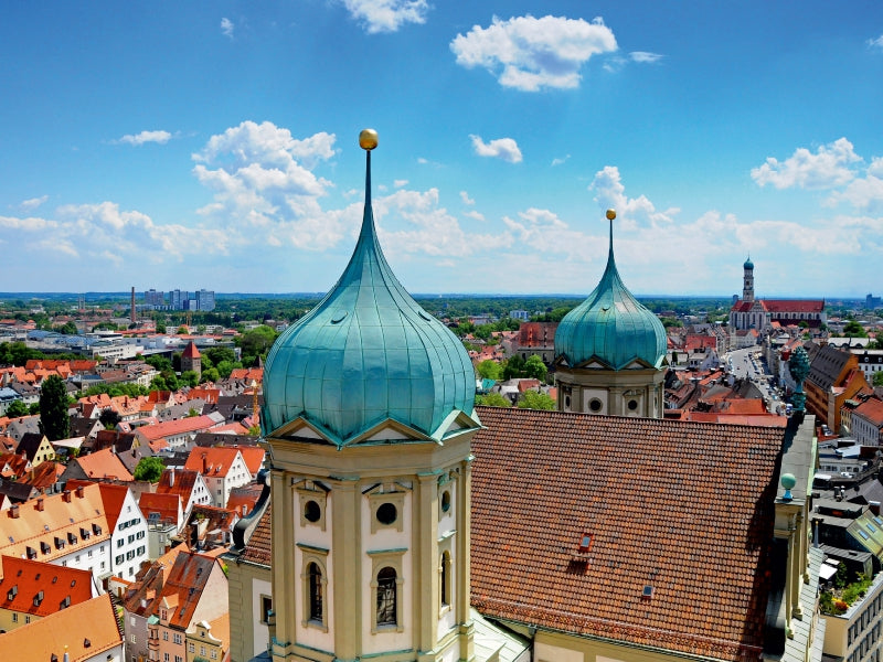 Blick von oben auf zwei grüne Kirchturmkuppeln und das rote Ziegeldach einer Stadt an einem sonnigen Tag mit Wolken.