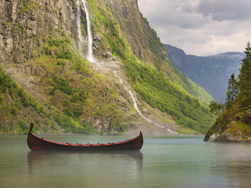 Ein Wikingerschiff schwimmt auf einem ruhigen Fjord vor steilen, bewaldeten Bergen mit Wasserfällen.