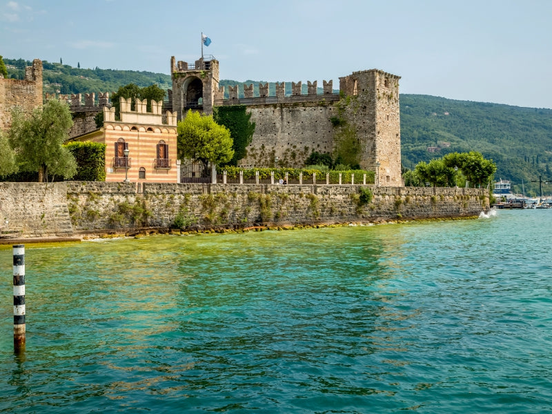 Ein mittelalterliches Schloss mit einer steinernen Mauer steht direkt am Ufer eines blauen Sees mit grünen Bergen im Hintergrund.