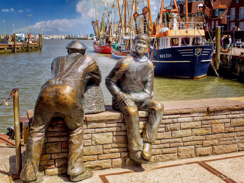 Bronzestatuen von zwei Fischern an einer Hafenmauer mit Booten im Hintergrund.