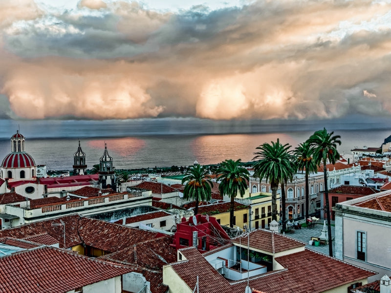 Blick über rote Ziegeldächer einer Stadt auf das Meer mit dramatischen Wolken am Himmel und Palmen.