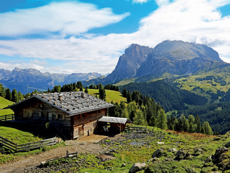 Eine rustikale Holzhütte auf einer grünen Alm vor einer imposanten Bergkette unter blauem Himmel.