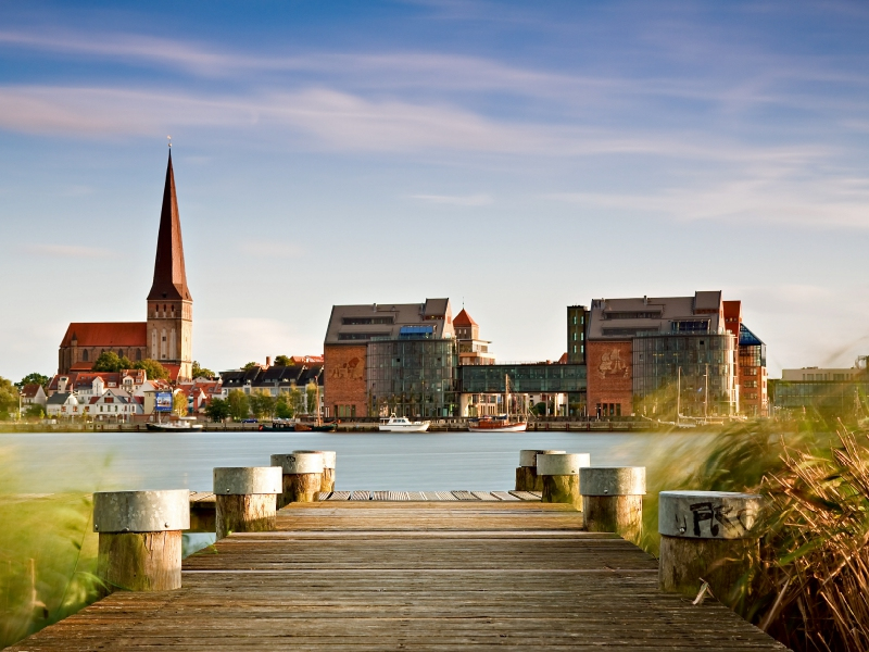 Holzsteg am Wasser führt zu einer Küstenstadt mit hoher Kirchturmspitze und modernen Gebäuden.