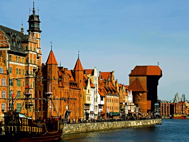 Historische Backsteingebäude und ein altes Segelschiff am Flussufer von Danzig unter blauem Himmel.