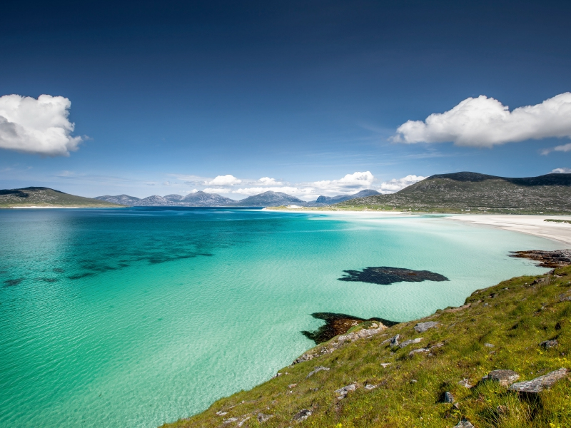 Türkisblaues Wasser und ein weißer Sandstrand unter blauem Himmel mit weißen Wolken.