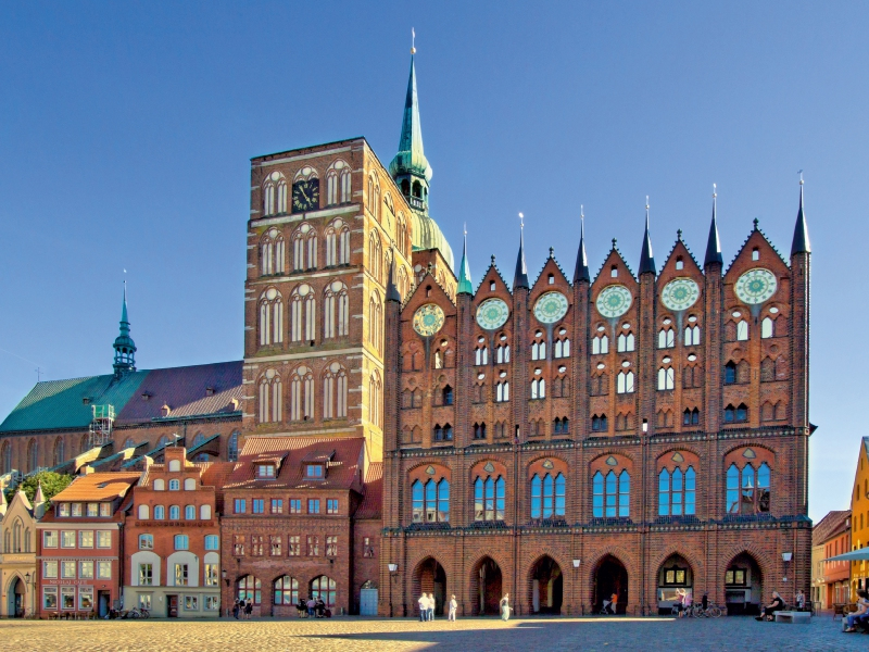 Historisches gotisches Rathaus aus roten Ziegelsteinen mit markanten Giebeln und Kirchturm im Hintergrund.
