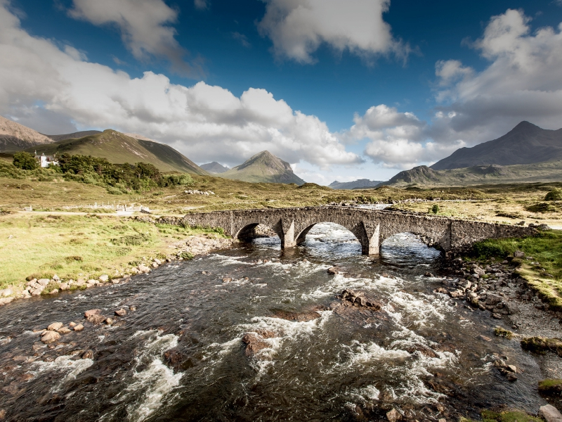 Alte Steinbrücke über einen rauschenden Fluss in einer bergigen Landschaft unter bewölktem Himmel.