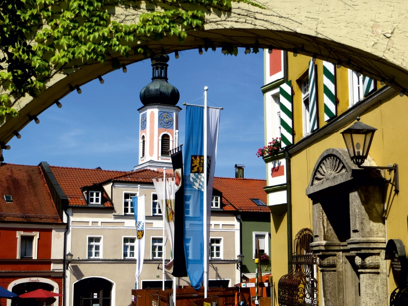 Blick durch einen Bogengang auf historische Gebäude, eine Kirchturmuhr und blau-weiße Flaggen unter klarem Himmel.