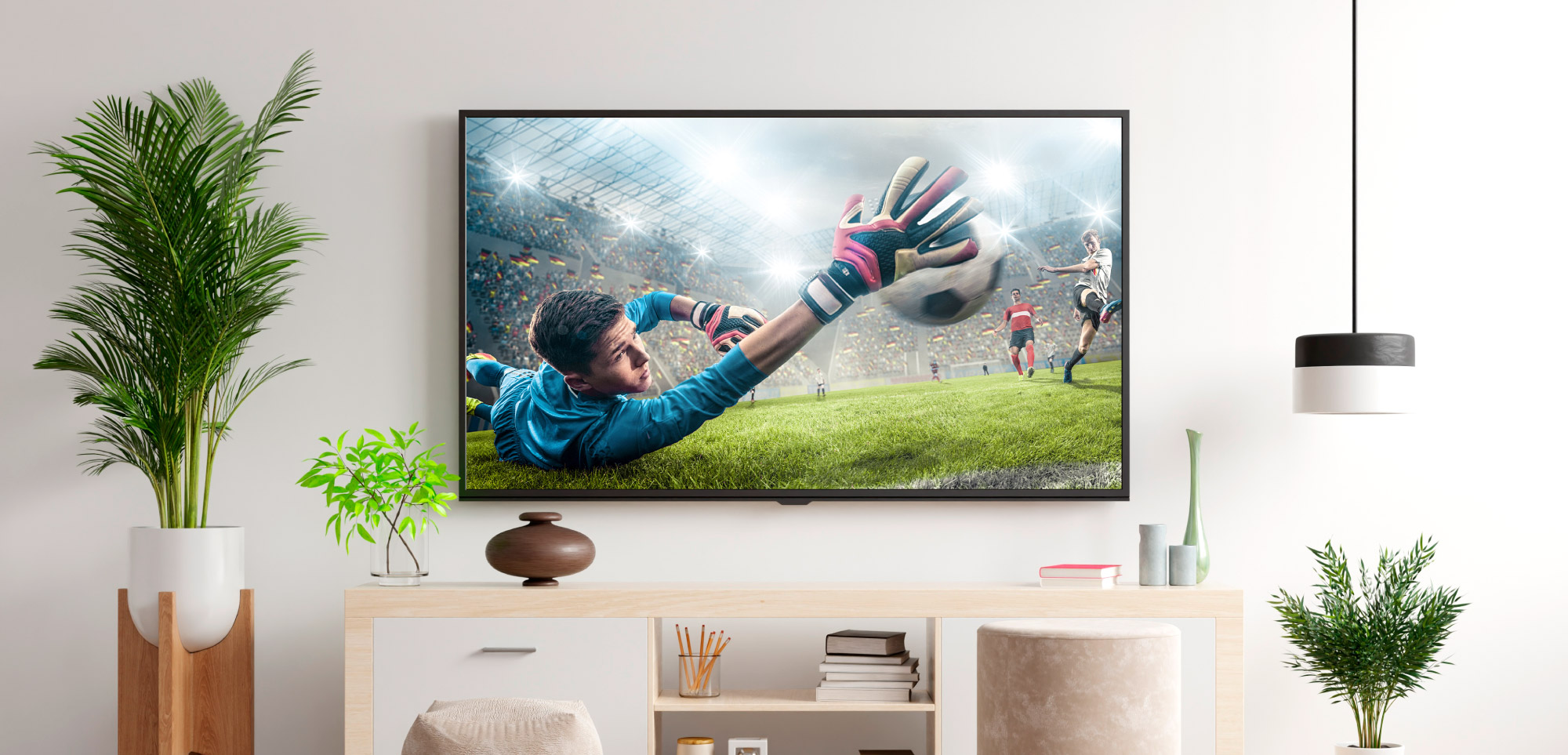 Telewizor pokazuje mecz piłki nożnej nad komodą, otoczony roślinami i lampą wiszącą.