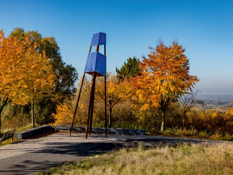 Blaue moderne Skulptur auf schlanken Beinen in herbstlicher Parklandschaft mit Bäumen in Gelb- und Orangetönen.