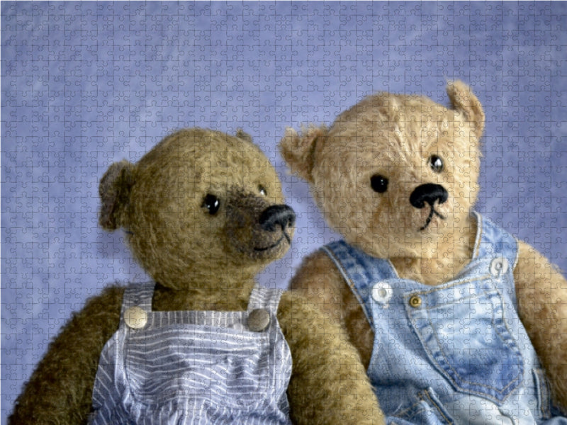 Zwei Teddybären in Jeans-Latzhosen sitzen nebeneinander vor blauem Puzzleteile-Hintergrund.
