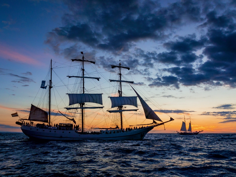 Ein großes Segelschiff segelt bei Sonnenuntergang auf ruhigem Meer unter einem wolkenverhangenen Himmel.