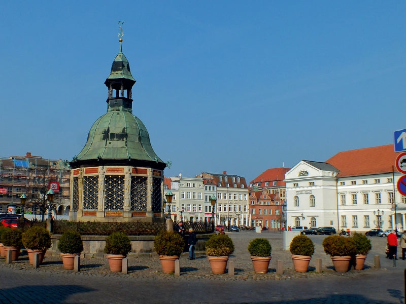 Ein historischer Brunnen mit grünem Kupferdach steht auf einem sonnigen Platz, umgeben von Topfpflanzen und Gebäuden.