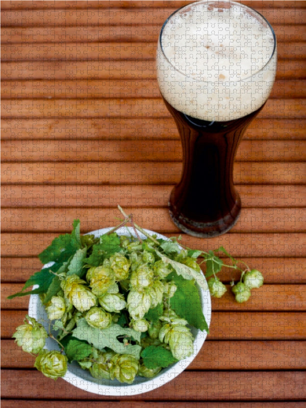 Ein Glas dunkles Bier mit Schaum und eine Schale mit frischem grünem Hopfen auf einem Holztisch.