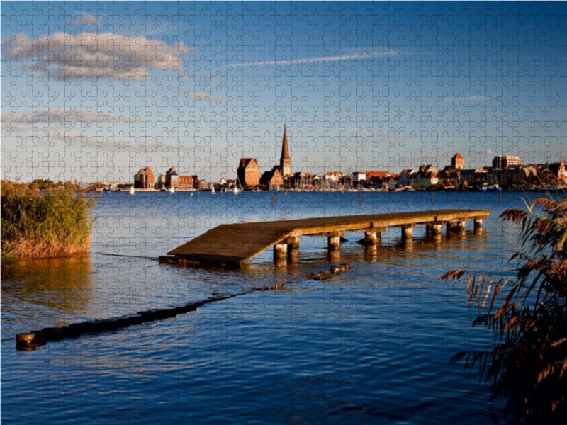Ein Holzsteg ragt in eine blaue Bucht, mit einer fernen Stadt und Kirchturm unter blauem Himmel.