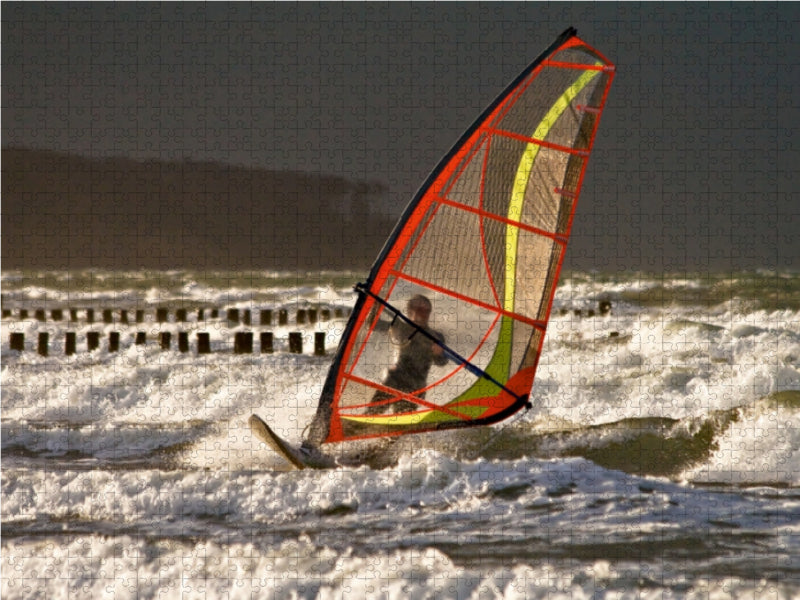 Ein Windsurfer mit einem orange-schwarzen Segel surft auf rauer See vor einer Küste.