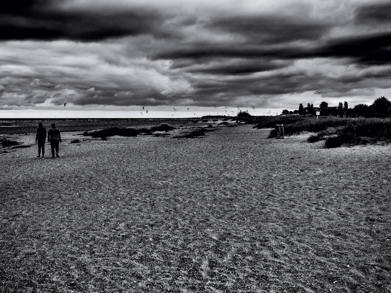 Zwei Personen gehen unter einem dramatischen, bewölkten Himmel an einem sandigen Strand entlang.