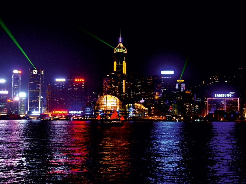 Beleuchtete Skyline einer Stadt bei Nacht mit Spiegelungen im Wasser und grünen Lasern.