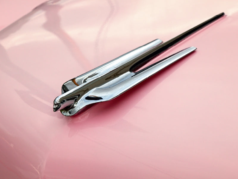 Eine verchromte Kühlerfigur in Form eines fliegenden Adlers auf der rosa Motorhaube eines Autos.