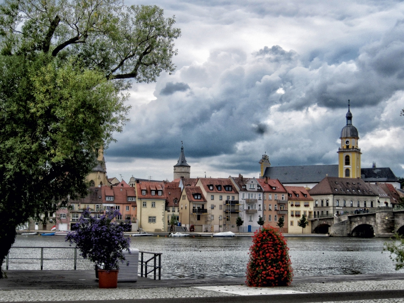 Uferblick auf eine historische europäische Stadt mit bunten Gebäuden, einer Brücke und bewölktem Himmel.