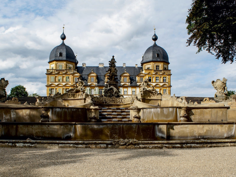 Barockschloss Weißenstein mit aufwendig gestaltetem Springbrunnen im Vordergrund und bewölktem Himmel.