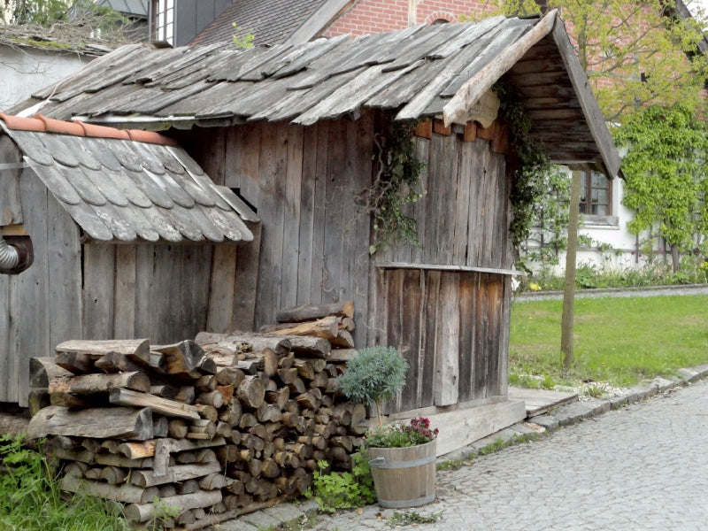 Eine alte Holzhütte mit Holzdach und Holzstapel davor auf einem Kopfsteinpflasterweg.