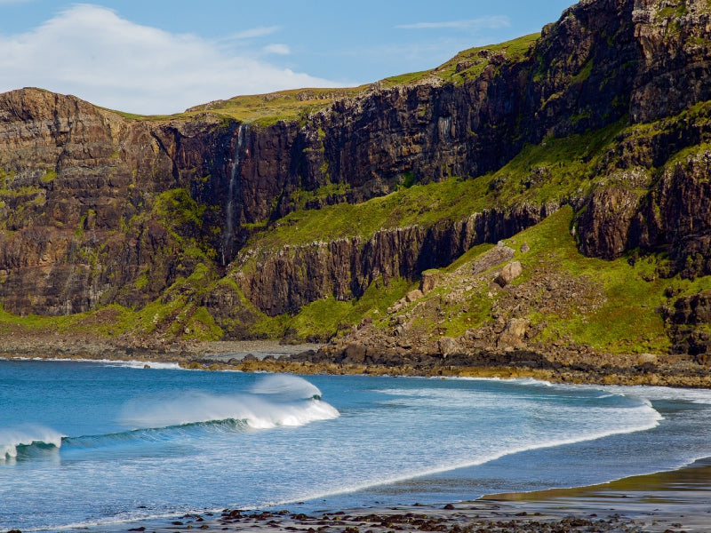 Grüne Klippen und ein kleiner Wasserfall treffen auf einen blauen Strand mit sanften Wellen.