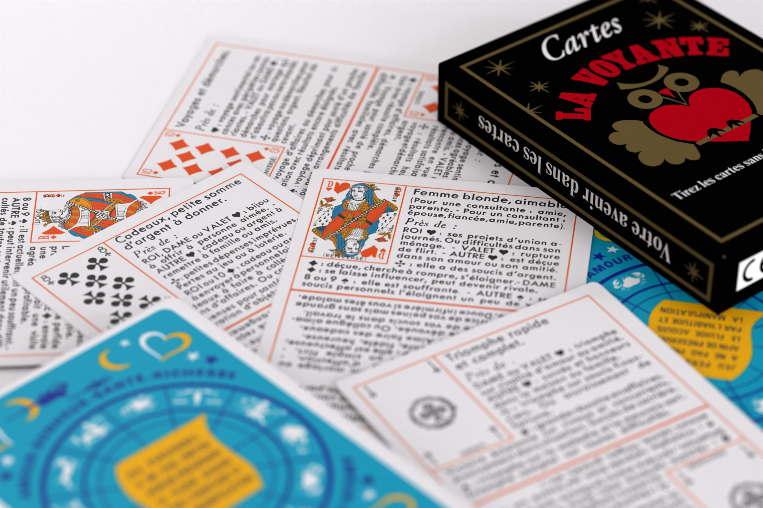 Französische Tarotkarten mit Text und Symbolen liegen neben einer schwarzen Kartenschachtel auf weißer Oberfläche.