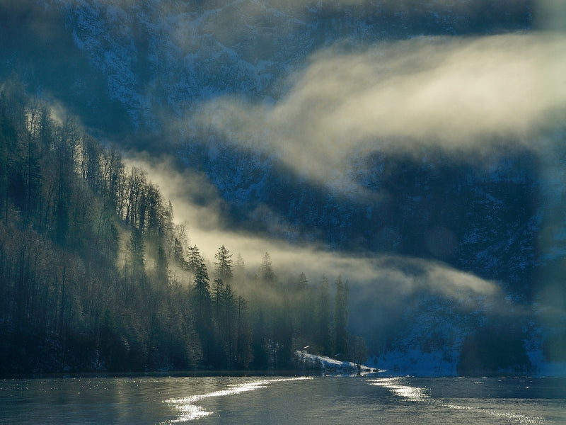 Nebel zieht über einen See und bewaldete Berge mit Schnee.