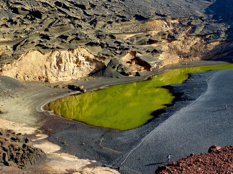 Eine leuchtend grüne Lagune zwischen dunklem Vulkanstrand und schroffen braun-schwarzen Klippen bei hellem Sonnenlicht.