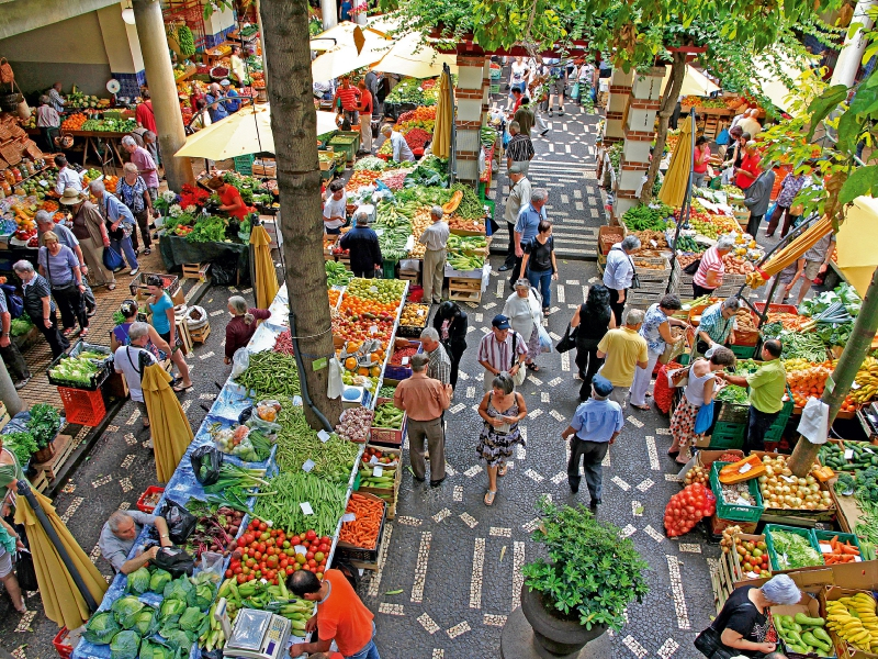 Belebter Markt mit vielen Ständen voller frischem Obst und Gemüse, umgeben von geschäftigen Menschen.