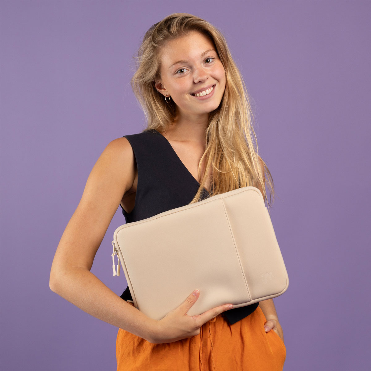 Mujer rubia sonriente con top negro y pantalones naranjas sostiene una funda de portátil beige ante un fondo morado.