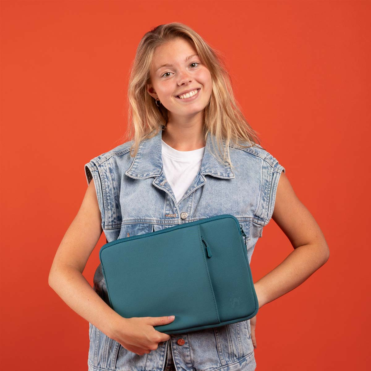 Una mujer rubia con chaleco de mezclilla sonríe sosteniendo una funda azul para laptop sobre fondo anaranjado.