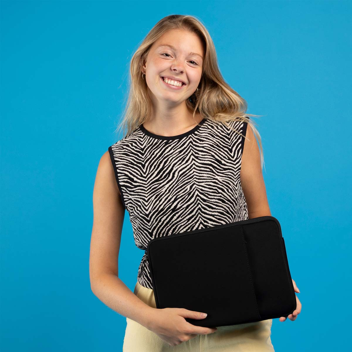 Una mujer rubia sonriente con top de cebra sostiene una funda de portátil negra ante fondo azul.