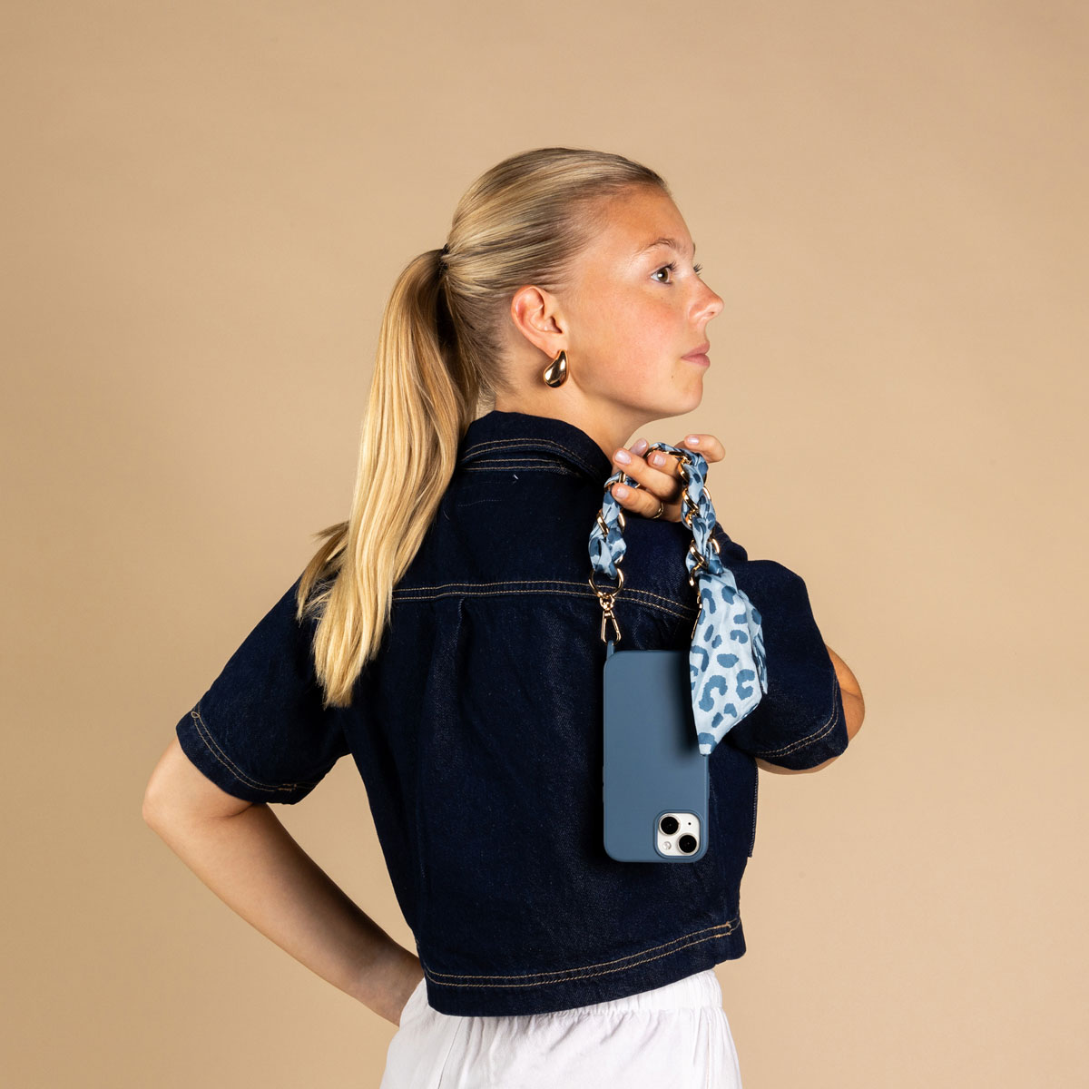 Eine blonde Frau im Denim-Hemd hält ein blaues Handy mit gemustertem Band auf beigem Hintergrund.