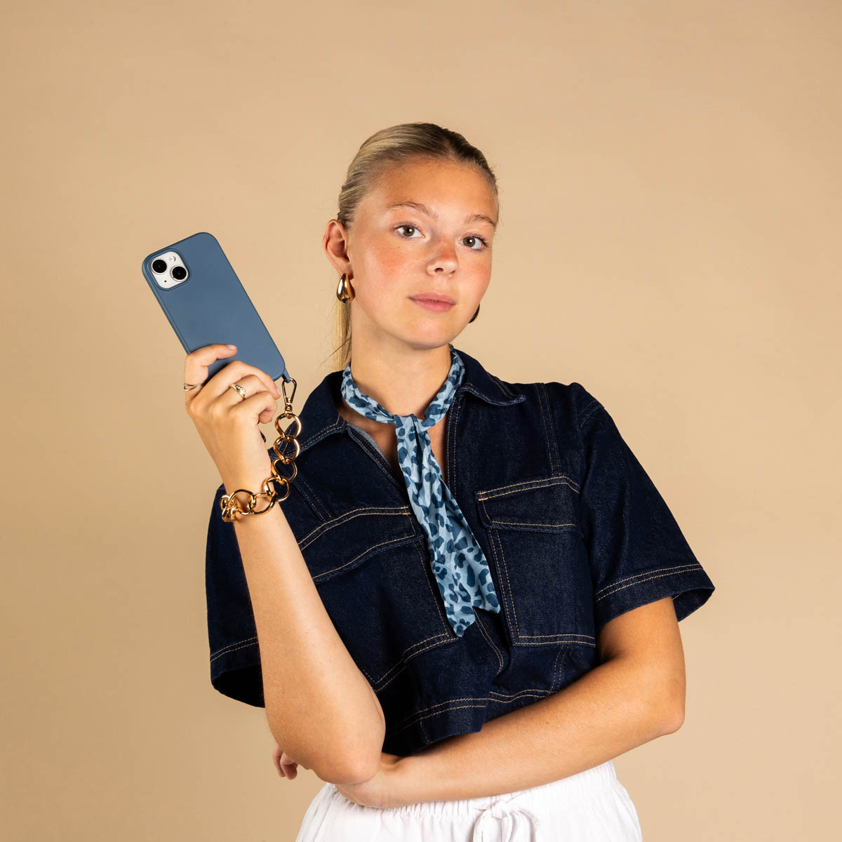 Eine blonde Frau im Jeanshemd hält ein blaues Smartphone mit einer goldenen Kette vor beigem Hintergrund.