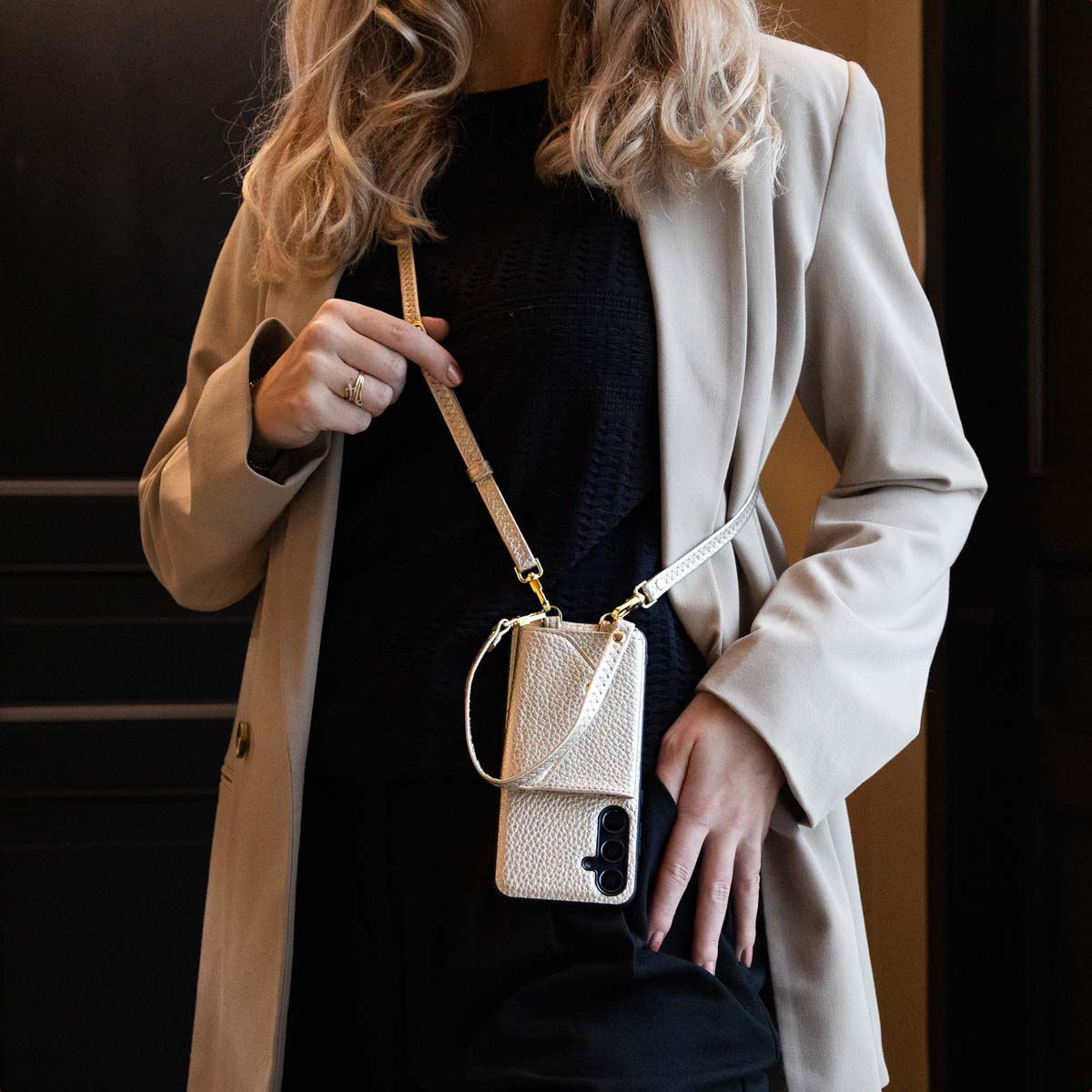 Une femme en blazer beige et haut noir porte un étui de téléphone doré avec une lanière sur fond sombre.